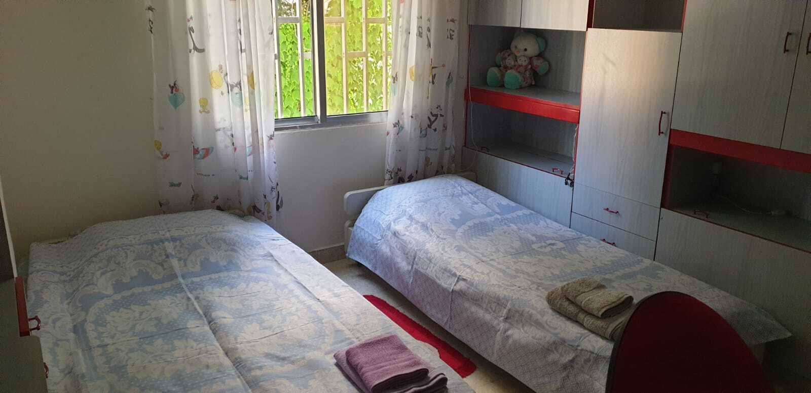 Ein Schlafzimmer mit zwei Betten, einem Fenster mit Vorhängen, einem Regal mit einem Teddybär und einem roten Stuhl.