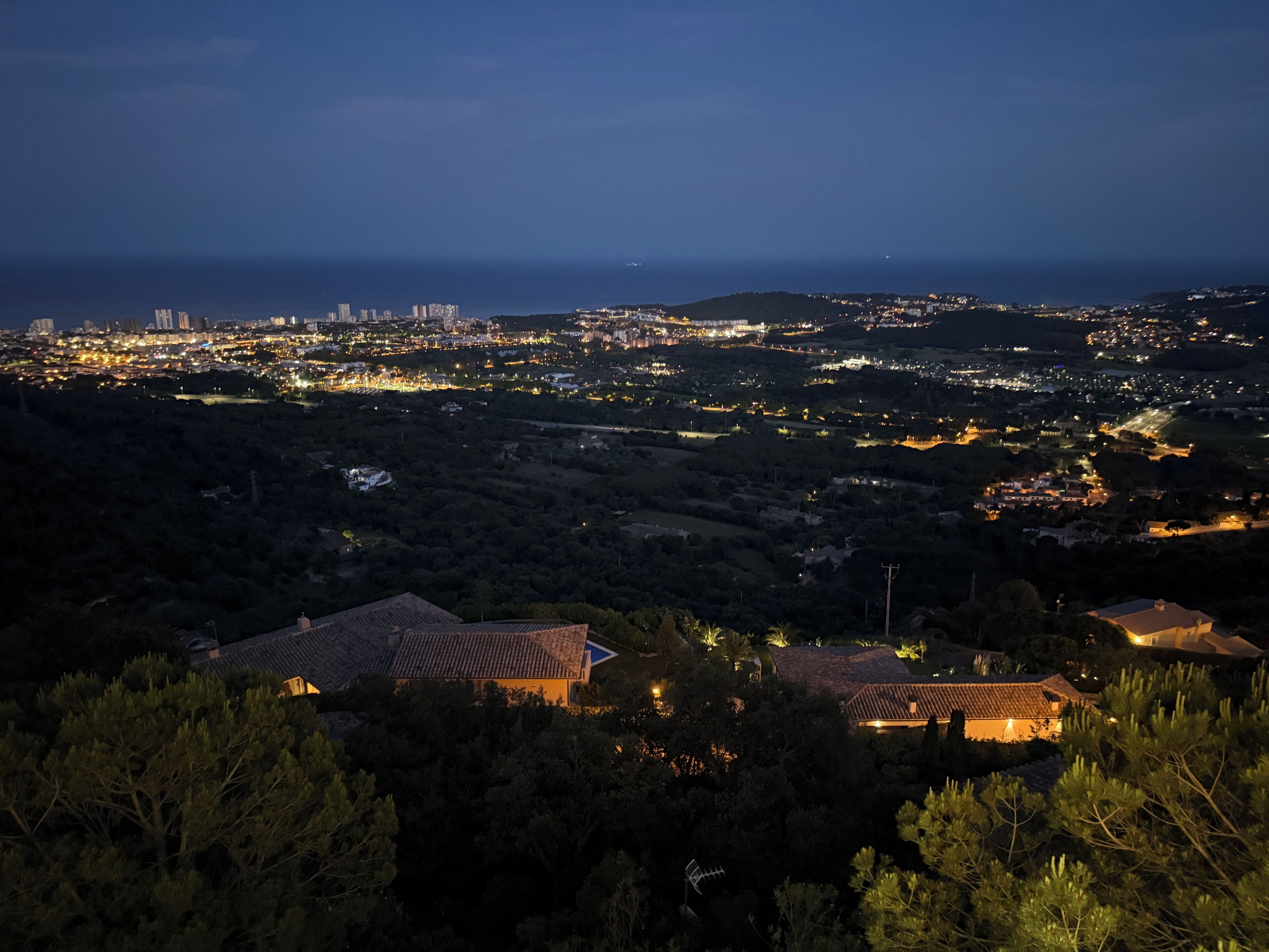 A panoramic view of a coastal city at night with illuminated buildings and a distant view of the sea.