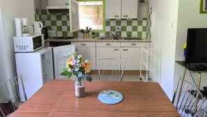 Et kompakt køkken med hvide hårde hvidevarer, et spisebord med en vase blomster og et fjernsyn på et stativ.