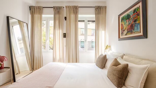 Une chambre d’hôtel avec un grand lit, des rideaux de couleur beige, un tableau au mur et une fenêtre donnant sur des immeubles.