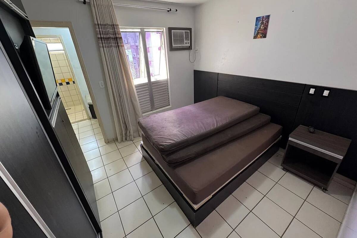 Ein Schlafzimmer mit einem Bett, einem Nachttisch, einem Fenster mit Vorhängen, einer Heizung und einer Wand-Klimaanlage.