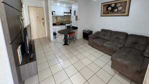 A living room with a brown sofa, a dining table with two chairs, a TV stand, and a kitchen in the background. - Eldorado Park (Caldas Novas)