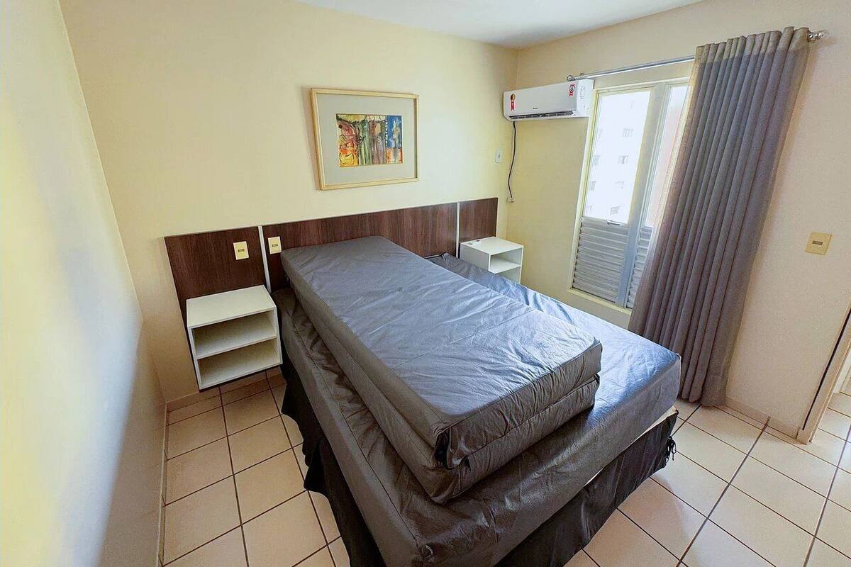 Ein Schlafzimmer mit Bett, Nachttisch, Wandklimaanlage, einem Fenster mit Vorhängen und einem gerahmten Bild an der Wand.