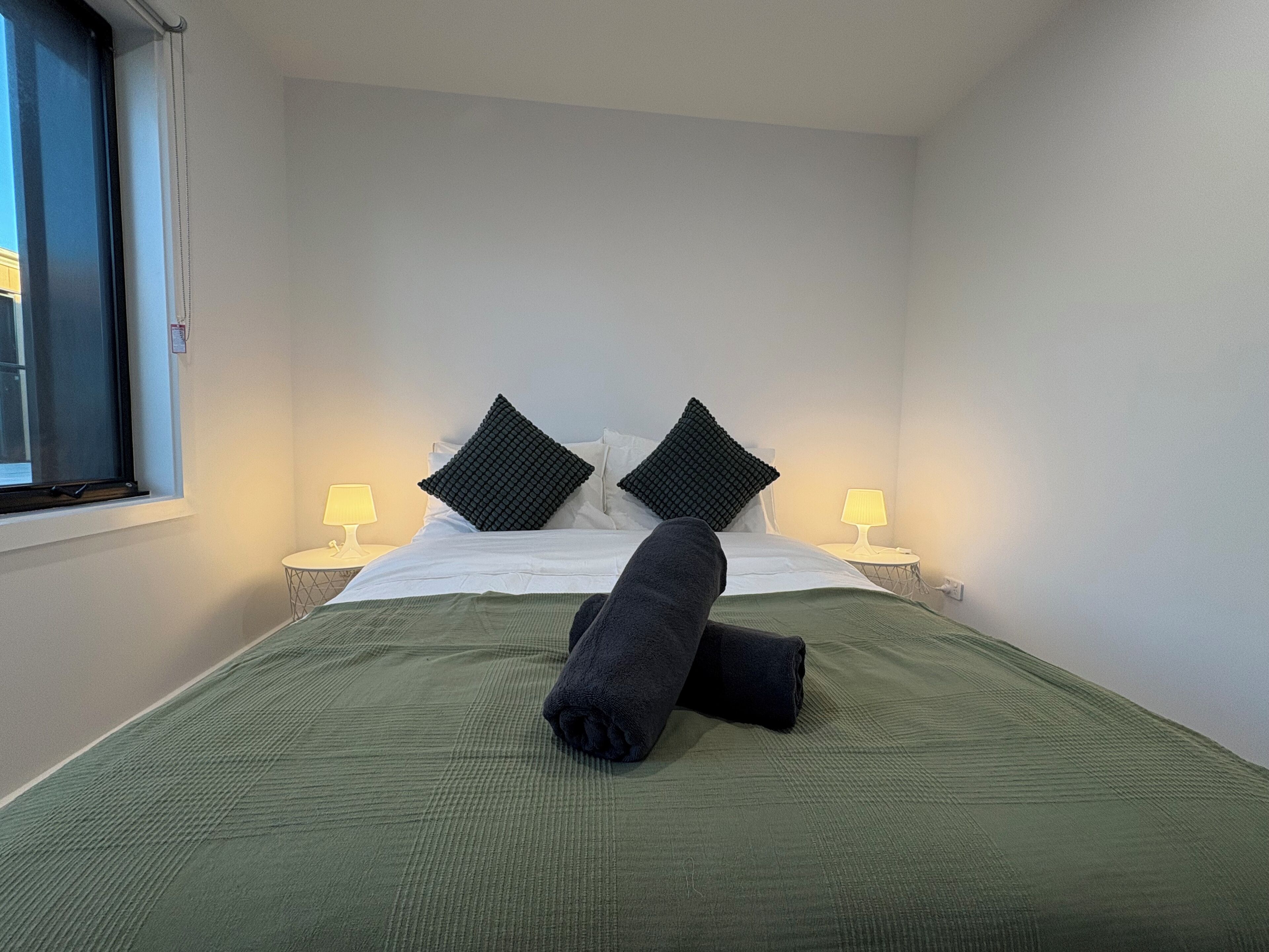A neatly made bed with a green bedspread and two black pillows, two bedside lamps, and a window with blinds.