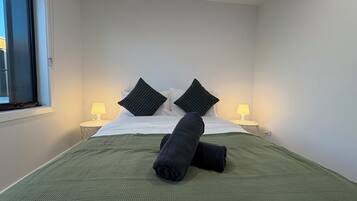 A neatly made bed with a green bedspread and two black pillows, two bedside lamps, and a window with blinds.