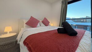 Habitación de hotel con cama, dos almohadas, una manta roja, una toalla negra, una mesita de noche con lámpara y ventana con vista a edificios.