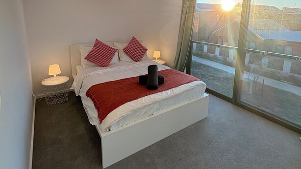 Un dormitorio moderno con una cama grande, una mesita de noche con una lámpara y vistas al exterior a través de un ventanal.