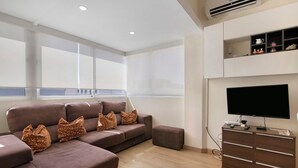 A modern living room with a sectional sofa, a flat-screen TV, a small cabinet, and a window providing natural light. - Charming Studio in Faro’s Historic Center – Faro Downtown (Faro)