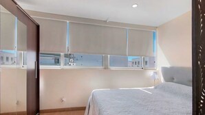 A bedroom with a bed, a window with blinds, a bedside lamp, and a view of buildings outside. - Charming Studio in Faro’s Historic Center – Faro Downtown (Faro)