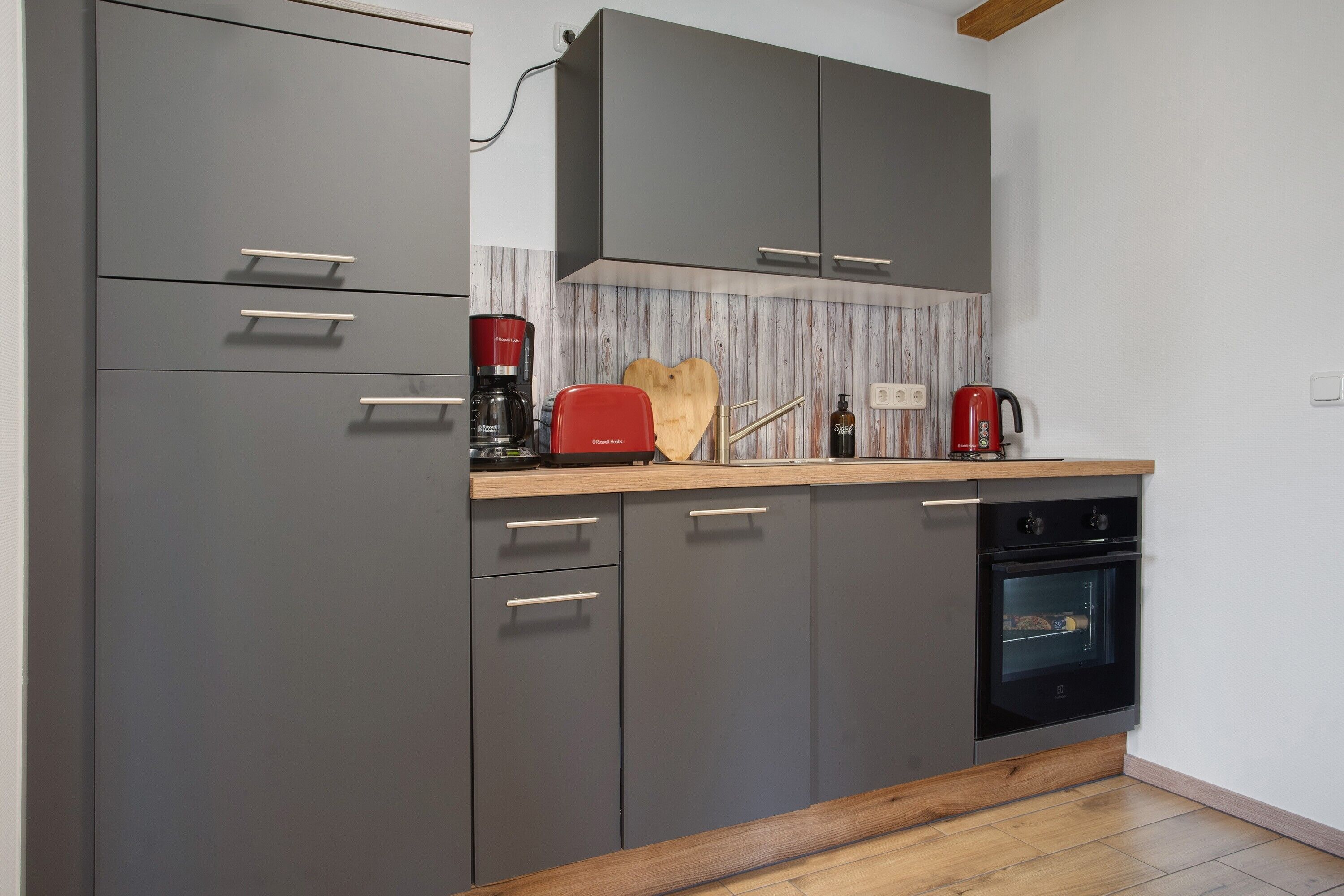 Une cuisine moderne avec des meubles gris, un plan de travail en bois, une machine à café rouge et un four encastré.