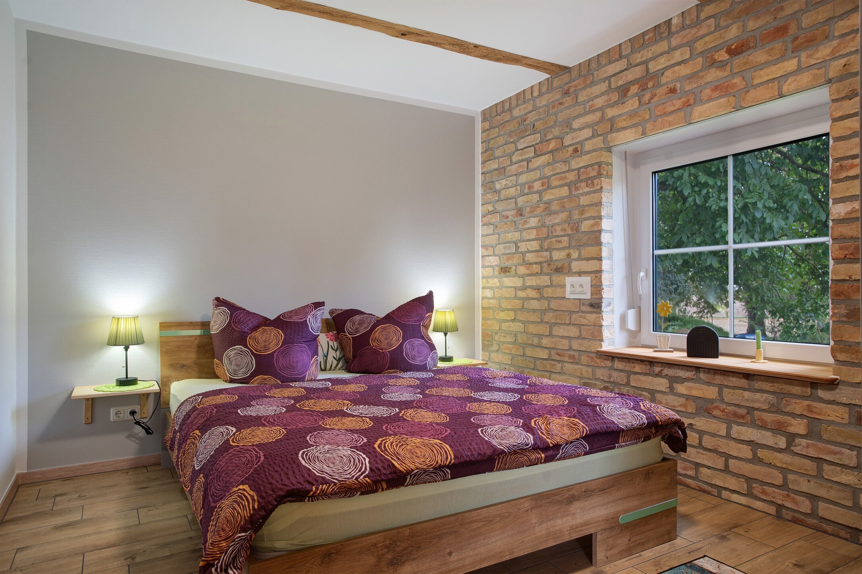 Ein Schlafzimmer mit Backsteinwand, einem Fenster, einem Bett mit gemusterten Betttextilien, zwei Nachttischlampen und einem Holz-Kopfstück.