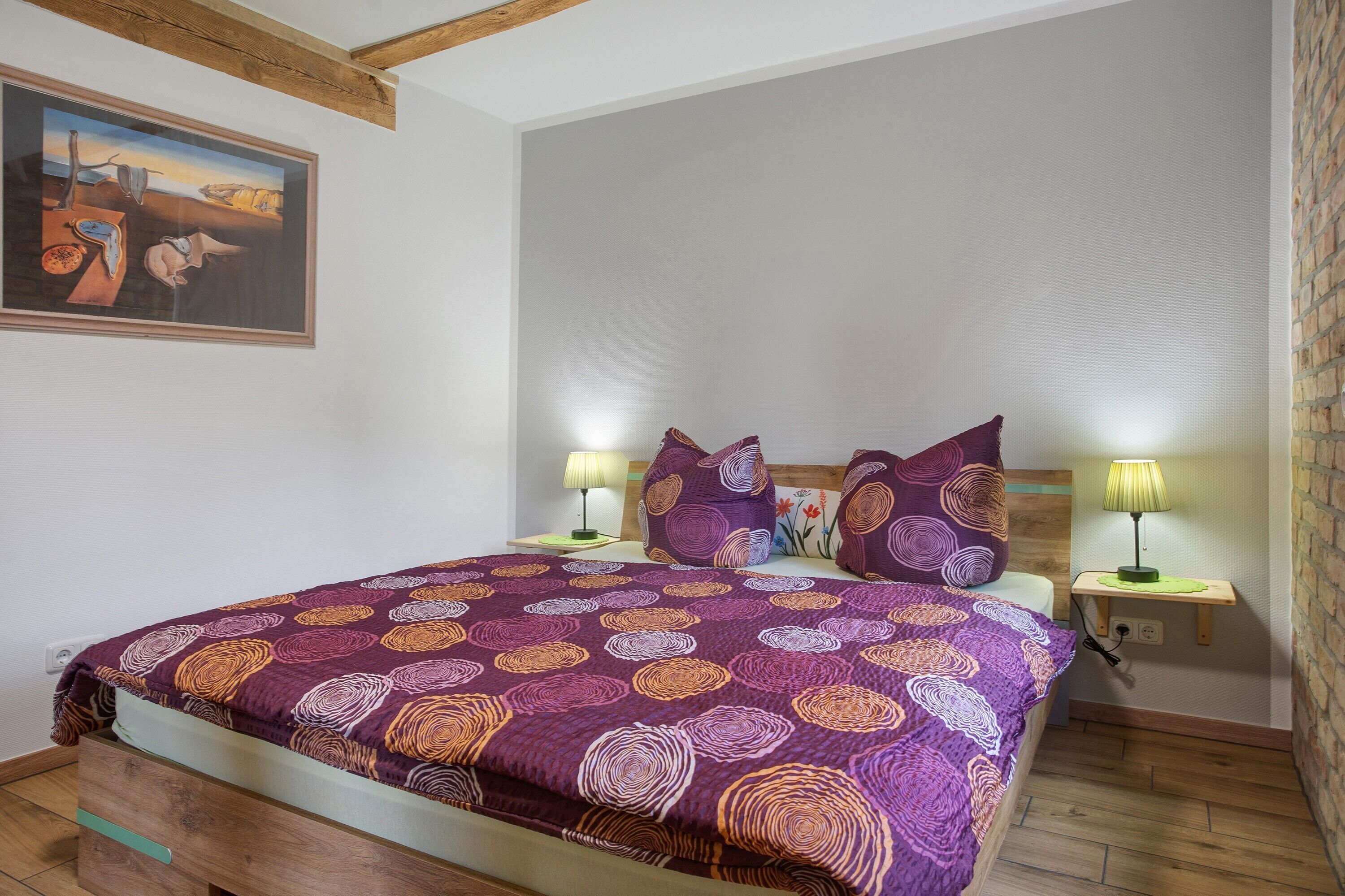 Un dormitorio con cama, dos lámparas de noche, un cuadro en la pared y suelo de madera.