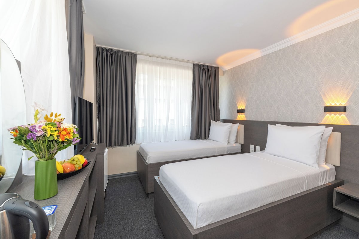 Quarto de hotel com duas camas, uma escrivaninha, um vaso de flores e uma fruteira.