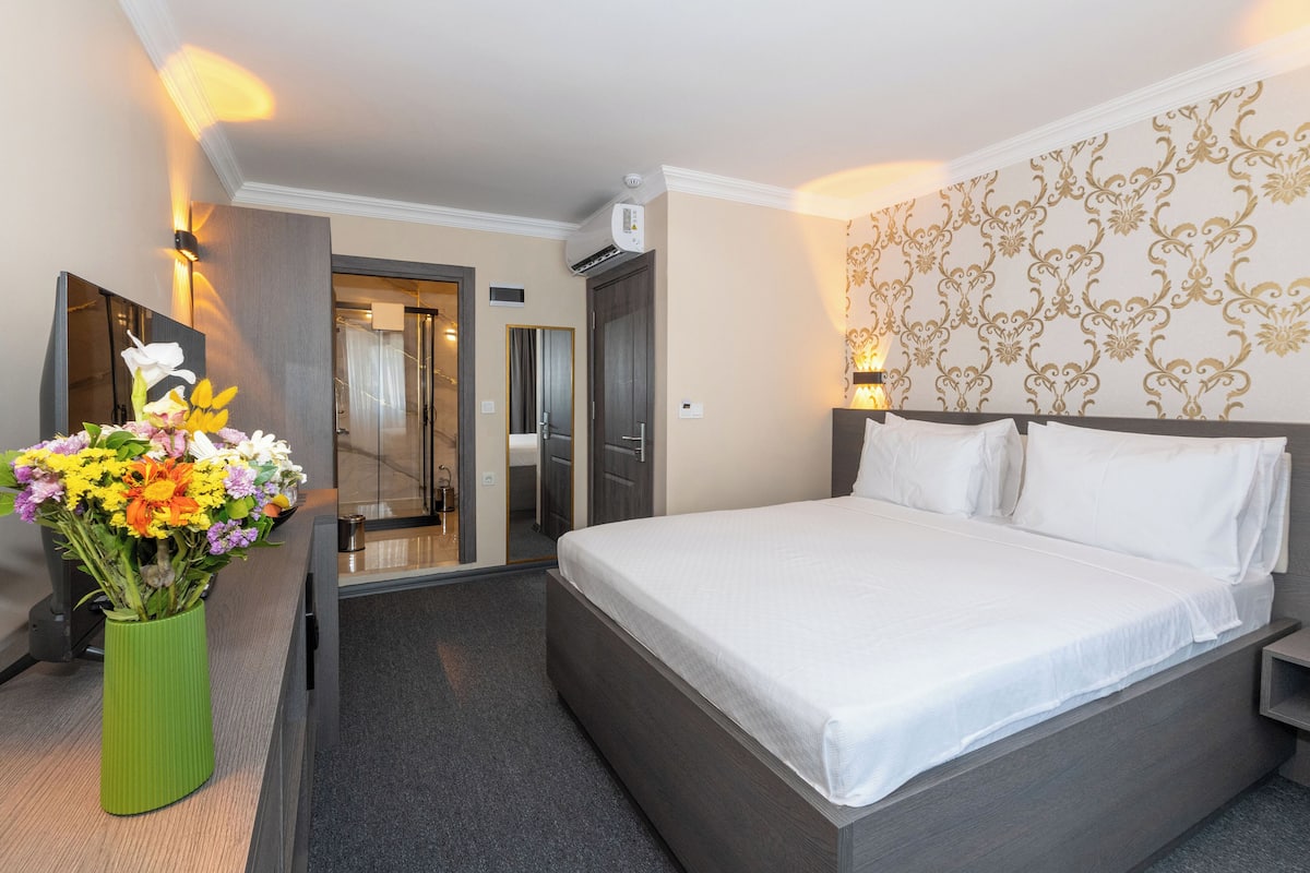 Quarto de hotel com uma cama grande, um vaso verde com flores, um espelho e parede estampada.