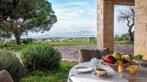 An outdoor dining area with a table set for breakfast, overlooking a scenic landscape with a view of the sea and mountains.