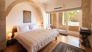 A bedroom with a large bed, a stone archway, a window with a view, and a small table with a lamp.