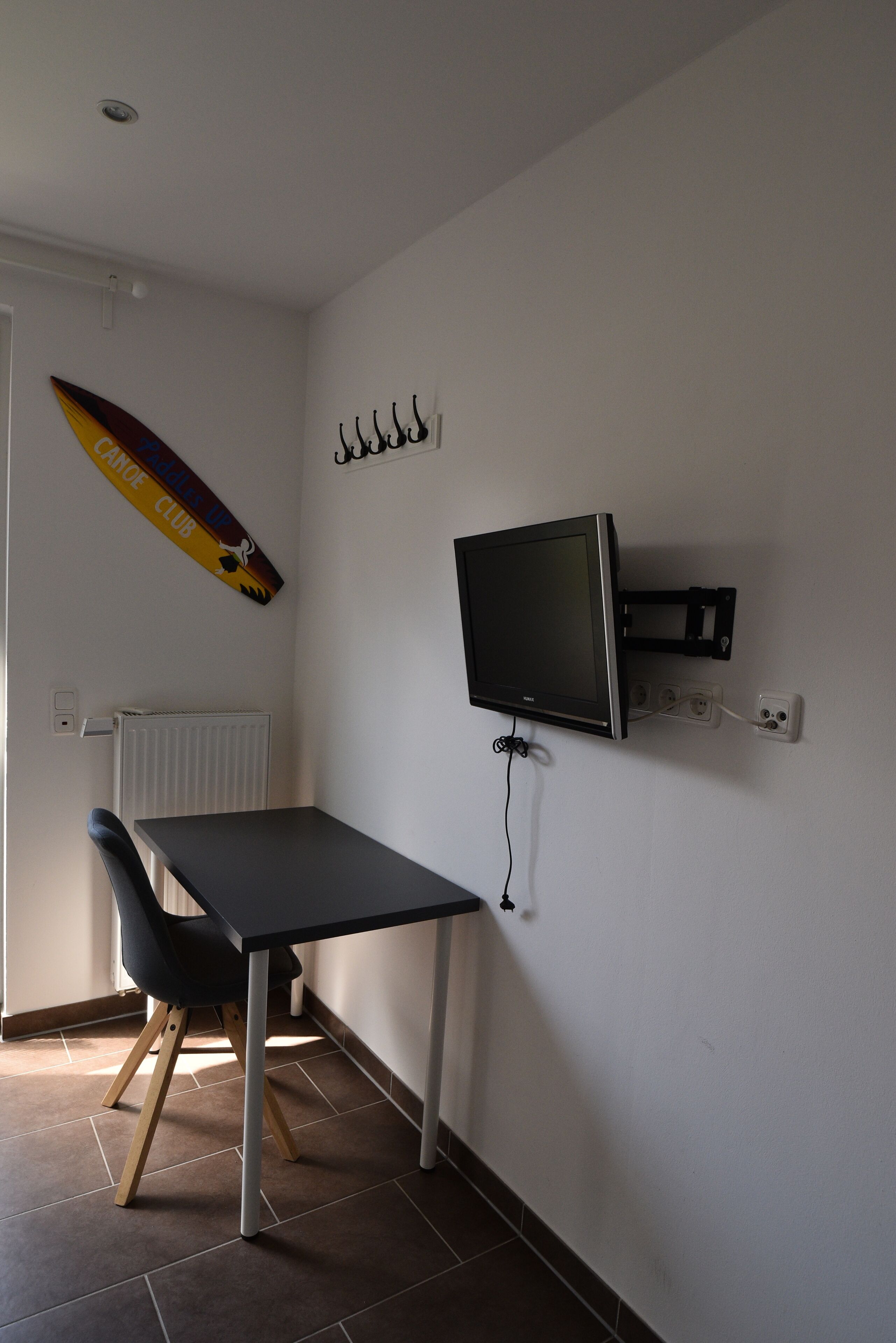 A room with a TV mounted on the wall, a table with a chair, and a surfboard decoration.