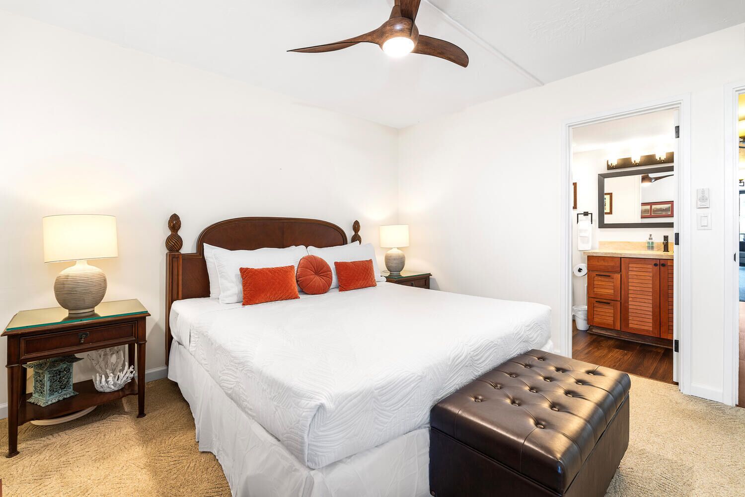 A bedroom with a large bed, two bedside tables with lamps, a wooden headboard, a ceiling fan, and a view into the bathroom.