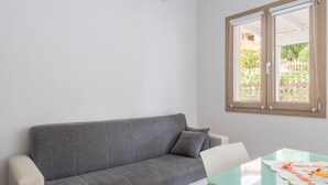 A grey sofa with a cushion, a white chair, a glass table with a patterned tablecloth, and a window with a view of greenery. - La Casa di Samu - White Apartment (Arbatax)