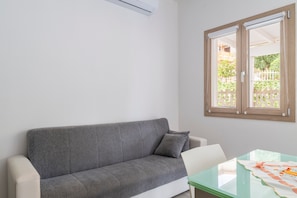 A grey sofa with a cushion, a white chair, a glass table with a patterned tablecloth, and a window with a view of greenery.