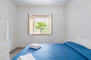 Quarto com cama, janela com persianas abertas e vista para o verde lá fora.