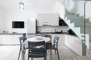 A modern kitchen with white cabinetry, a dining table with chairs, a TV mounted on the wall, and a staircase with glass railings.