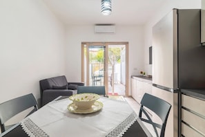 Een kleine keuken met een koelkast, een eettafel met stoelen en uitzicht door een glazen deur op een buitenruimte.