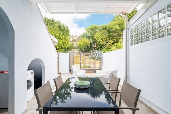 Un patio cubierto con una mesa, sillas, lavadora y vistas a árboles y una valla.