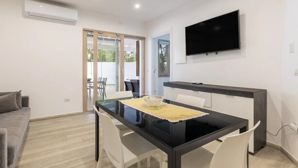 Un salon moderne avec une table à manger en verre, des chaises blanches, un canapé, un téléviseur à écran plat fixé au mur et un appareil de climatisation.