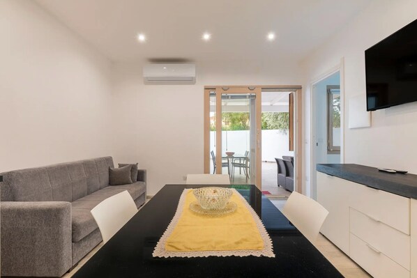 A modern dining area with a black table, white chairs, a grey sofa, and a TV on the wall.
