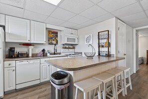 A modern kitchen with white cabinetry, a central island with stools, a microwave, and a trash can. - Coastal Retreat Steps From Private Beach 2 Bedroom Apts (Murrells Inlet)