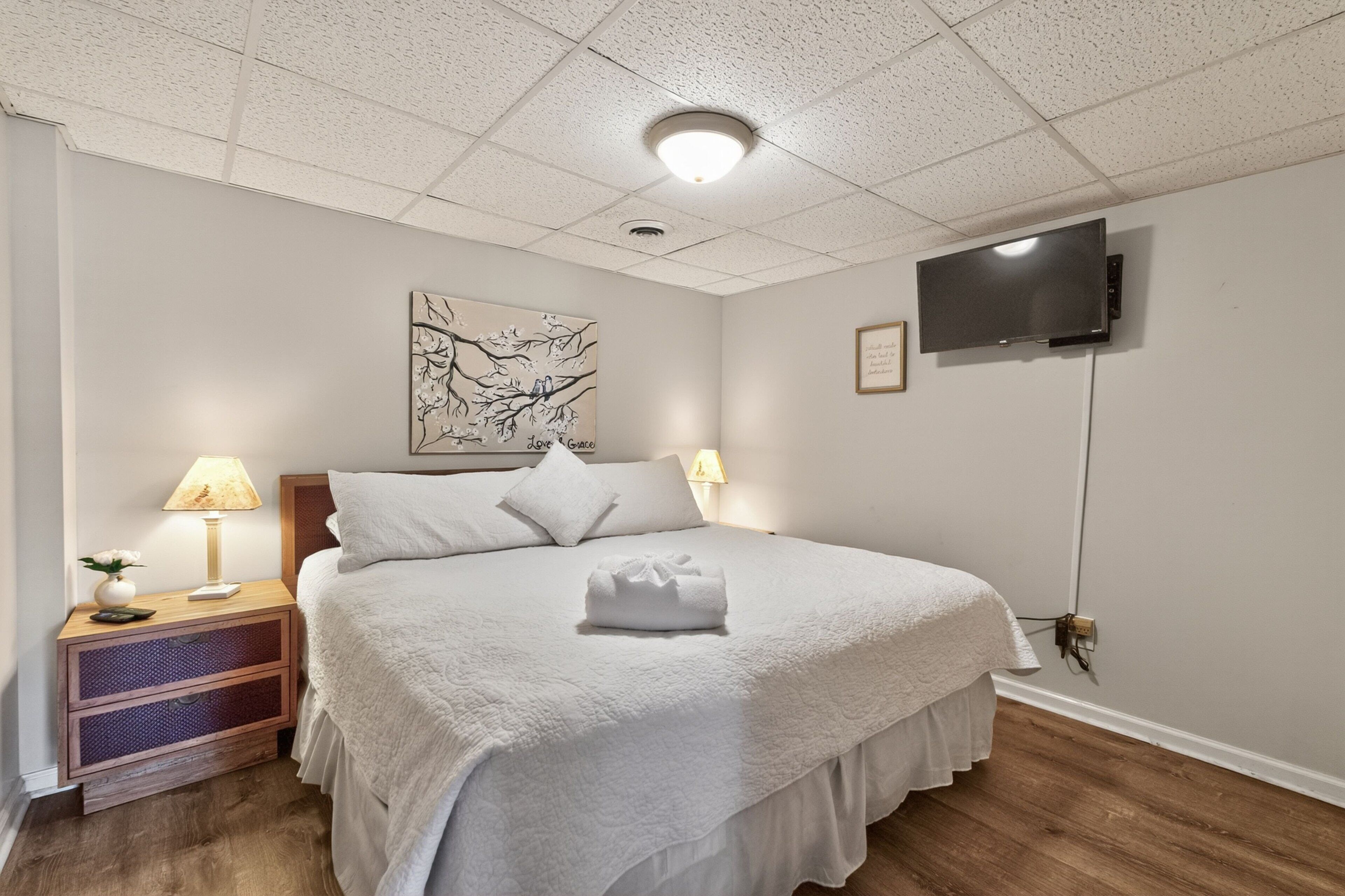 A neatly made bed with a white comforter and pillows, a bedside table with a lamp, a wall-mounted TV, and a framed artwork above the bed.