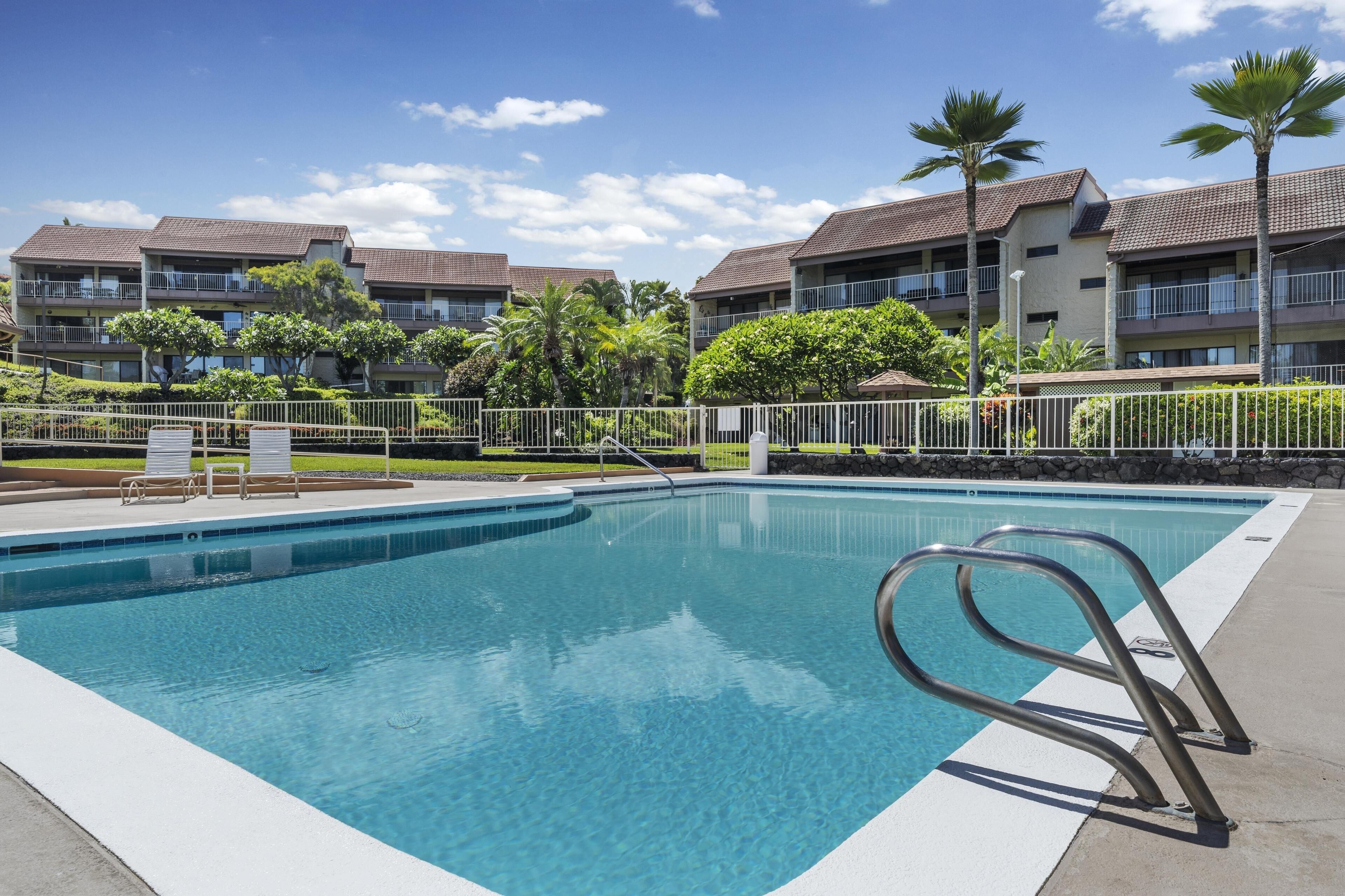 A swimming pool with a metal ladder, surrounded by a fence, and adjacent to a building with multiple balconies and palm trees.