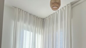 Une pièce dotée d’une grande fenêtre ornée de rideaux blancs et légers, d’un plancher en bois, d’un canapé marron et d’un petit tabouret en bois.