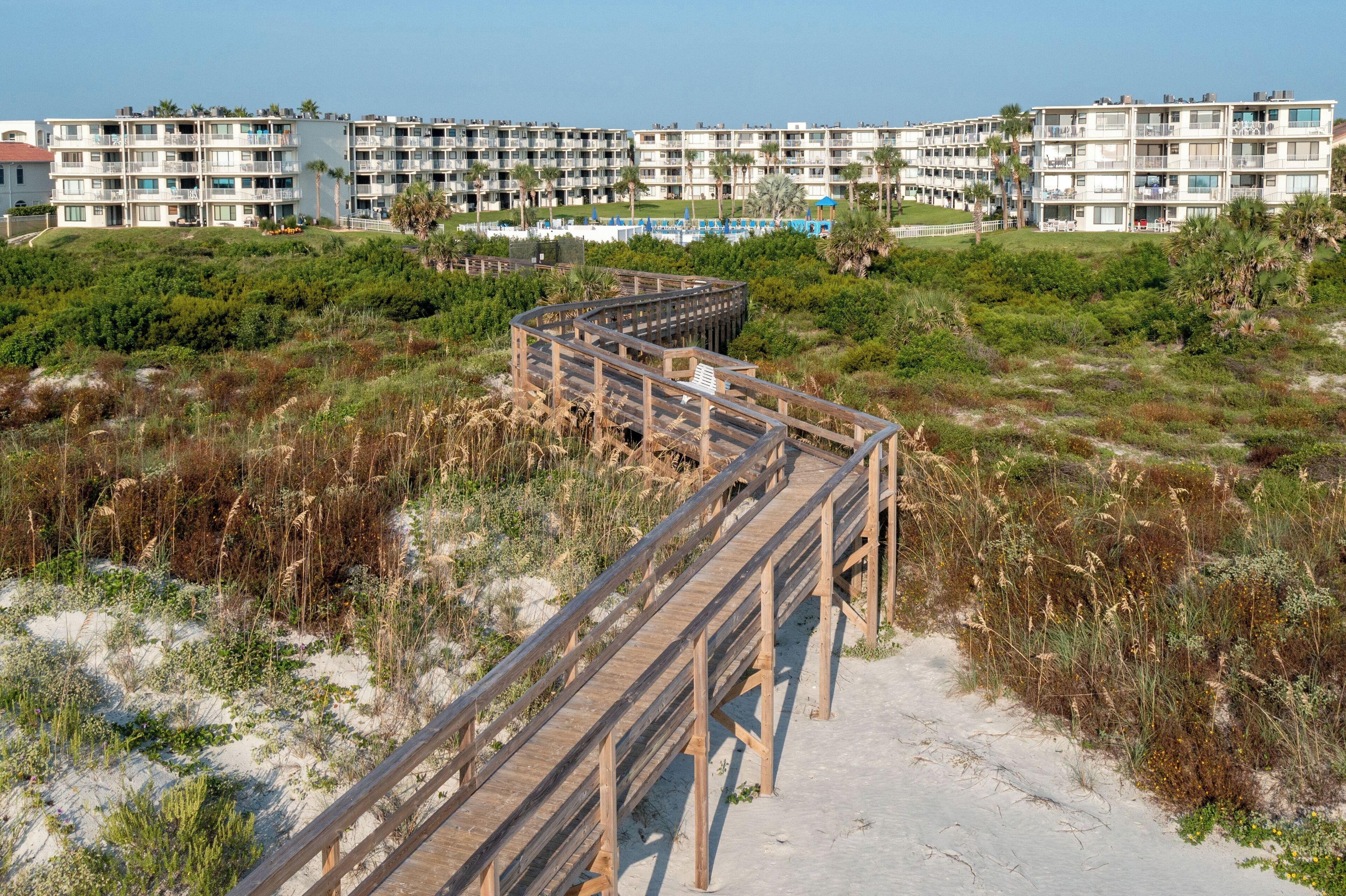 A wooden boardwalk leading to a beachfront hotel with multiple balconies and a pool area.