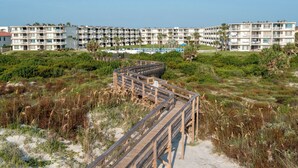 Une promenade en bois mène à un hôtel en bord de mer, doté de nombreux balcons et d’un espace piscine.