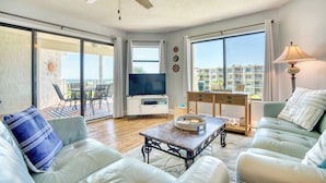 Un vaste salon doté d’un ventilateur de plafond, d’un téléviseur à écran plat, d’un coin repas avec une table et des chaises, et de grandes fenêtres offrant une vue sur la ville.