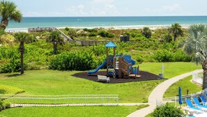 Une aire de jeux avec un toboggan et des structures d’escalade, entourée d’une zone herbeuse délimit ée par une clôture et d’un sentier menant à la plage.