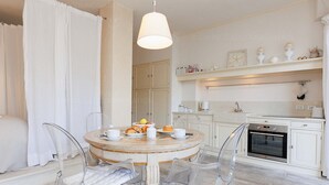 A dining area with a round table set for breakfast, clear chairs, a hanging light fixture, and a kitchen in the background. - CG Armenonville White-2811-very charming studio near the Croisette (Cannes)