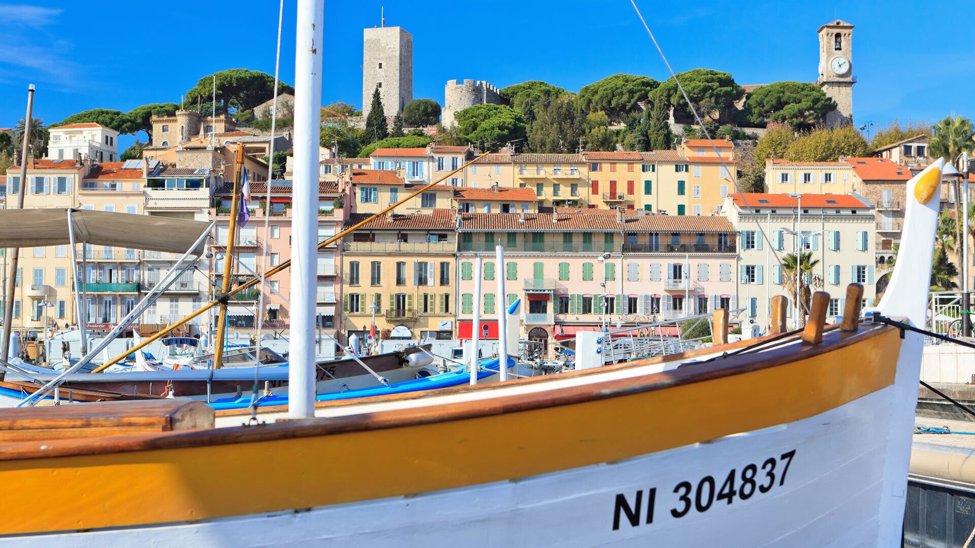 A boat with the number NI 304837 docked in a marina with colorful buildings and a tower in the background.