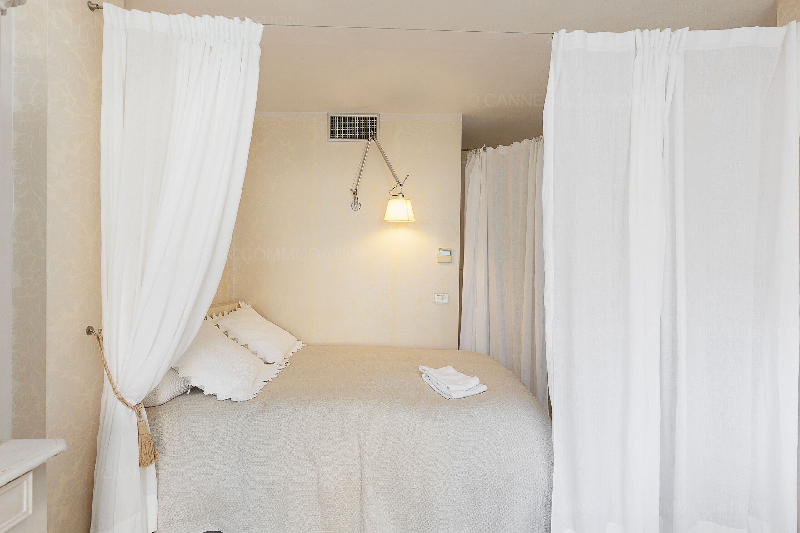A neatly made bed with white bedding, a hanging lamp, and sheer curtains.