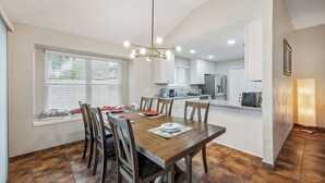 A dining area with a wooden table, chairs, and a chandelier. There is a window with blinds and a view of the outside. The kitchen is visible in the background with cabinets and appliances. - Space & Sunshine Retreat/3BR Near NASA & NRG (Houston)