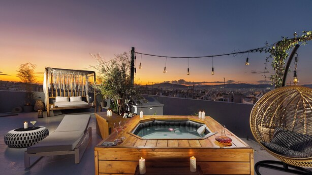 Eine Dachterrasse mit einem Holz-Hottub, Liegestühlen und Lichterketten vor einer Sonnenuntergangs-Stadtlandschaft.