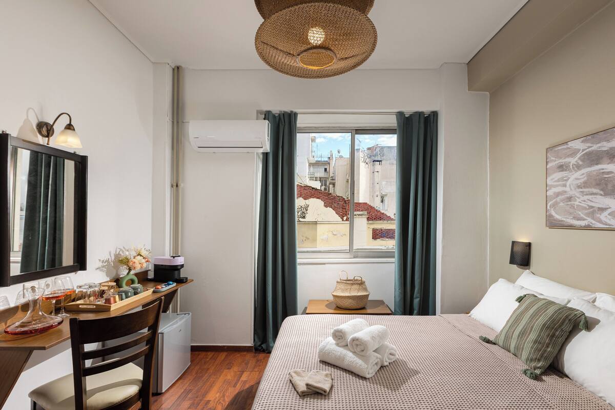 A hotel room with a bed, a desk, a chair, a window with curtains, and a view of buildings outside.