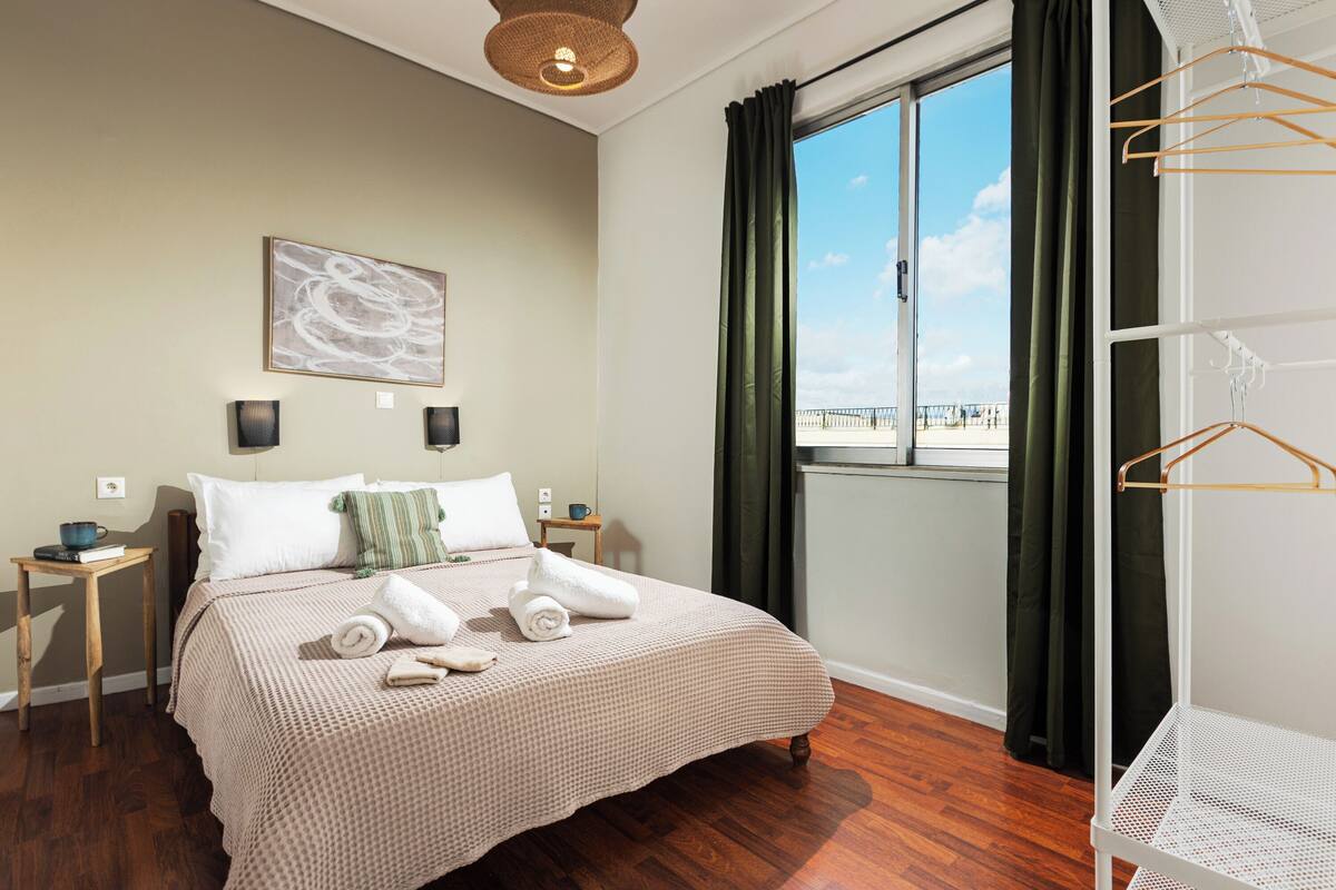 A neatly made bed with a textured bedspread, a bedside table with a cup and book, a wall-mounted artwork, and a view of a balcony through a window with curtains.