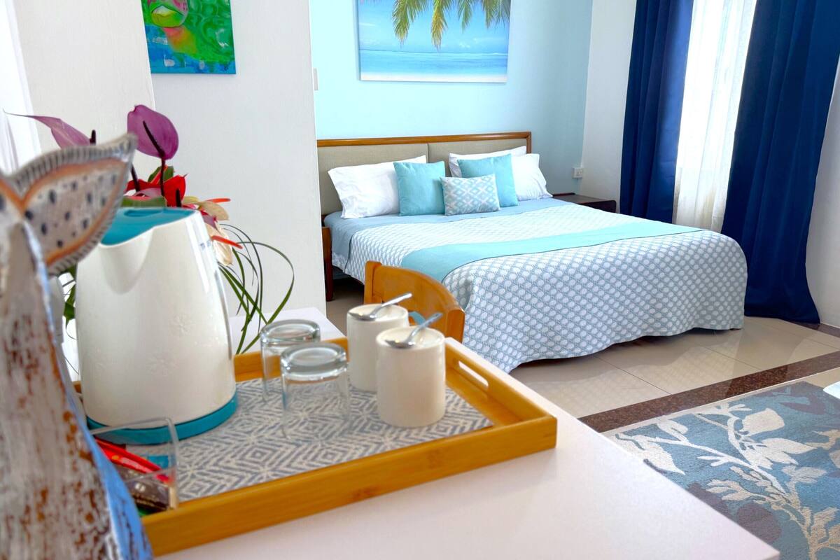 Une chambre d’hôtel avec un lit, un plateau contenant des articles pour le petit-déjeuner, un tableau représentant une plage et une fenêtre avec des rideaux.