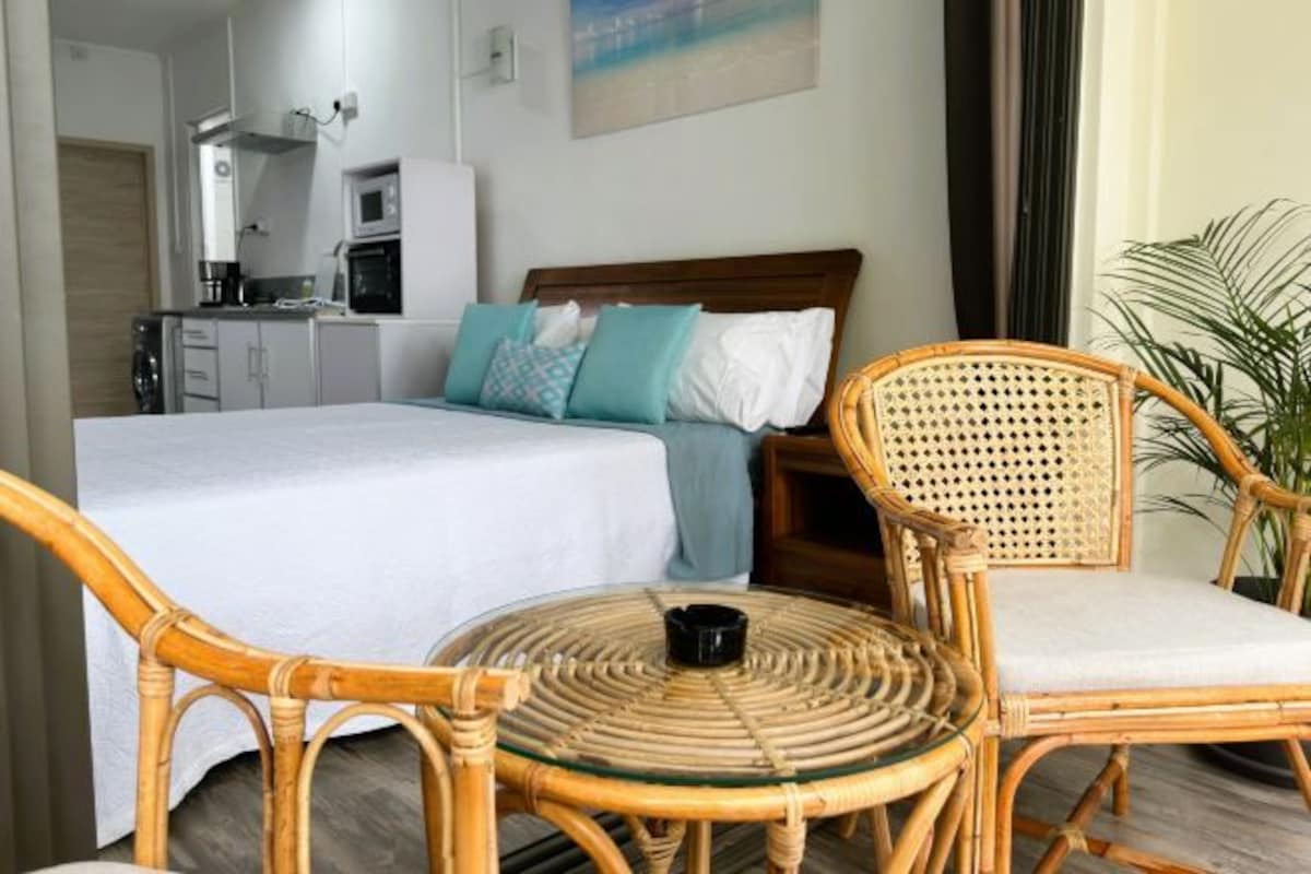 A hotel room with a bed, a small kitchenette, a round wicker table, and two wicker chairs.