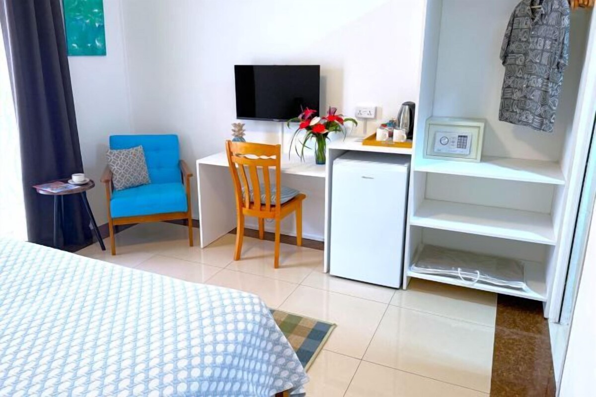 A room with a bed, a desk with a computer, a chair, a small fridge, and a shelf with items.