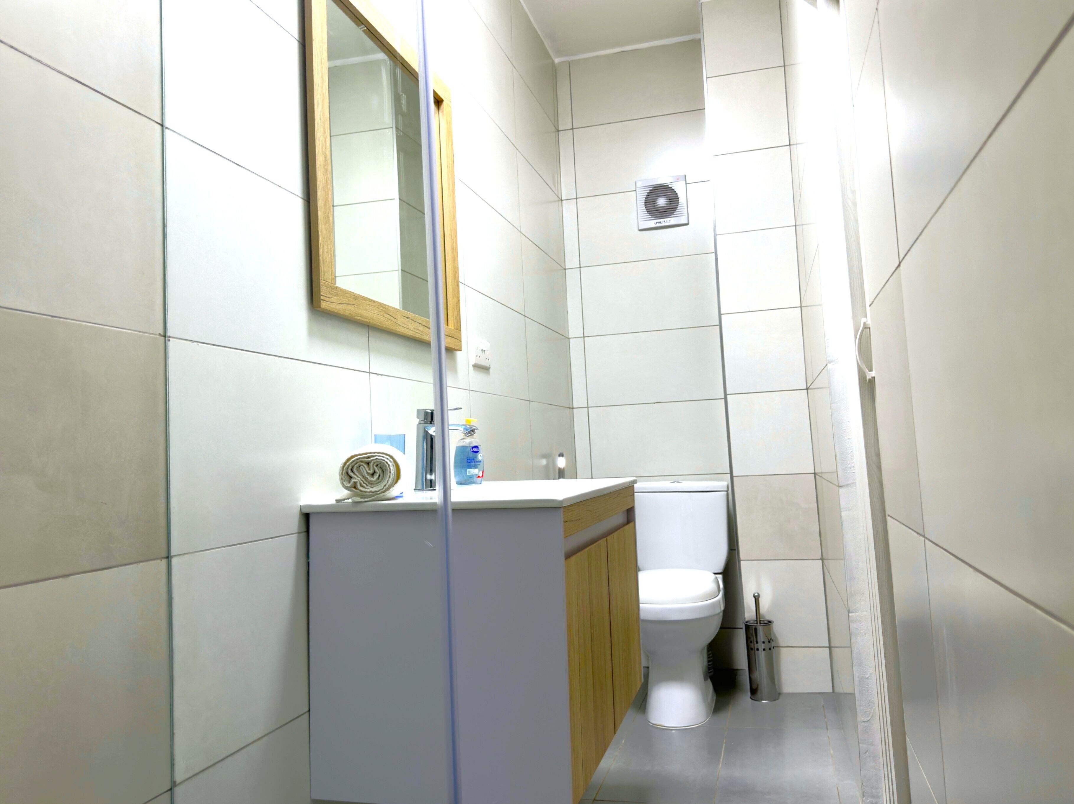 Un baño pequeño y moderno con azulejos blancos, un mueble de tocador de madera, un inodoro y un espejo.