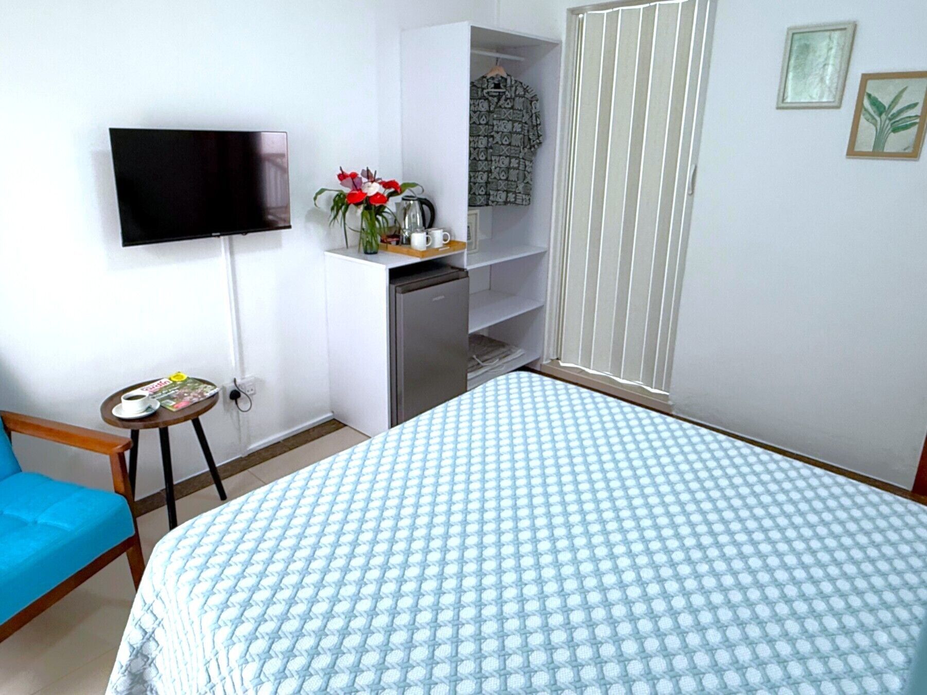 A bedroom with a bed, a TV mounted on the wall, a small table with a cup and magazine, a blue chair, and a kitchenette area.